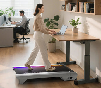 A woman walks on a treadmill desk while using a laptop in a bright office with plants and a colleague working nearby.