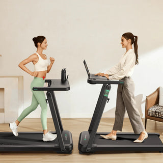 Two women use treadmills: one exercises in sportswear, while the other works on a laptop in casual attire.